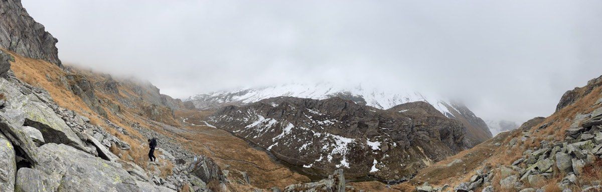 A hiker climbing above the open Greina plateau beneath low cloud and patchy snow